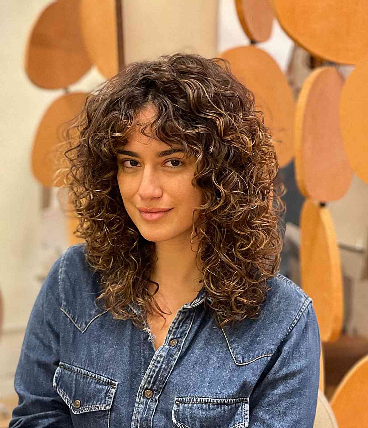 A woman with a medium-length Cadō cut featuring natural curls and face-framing curtain bangs, ideal for wavy to curly hair textures.