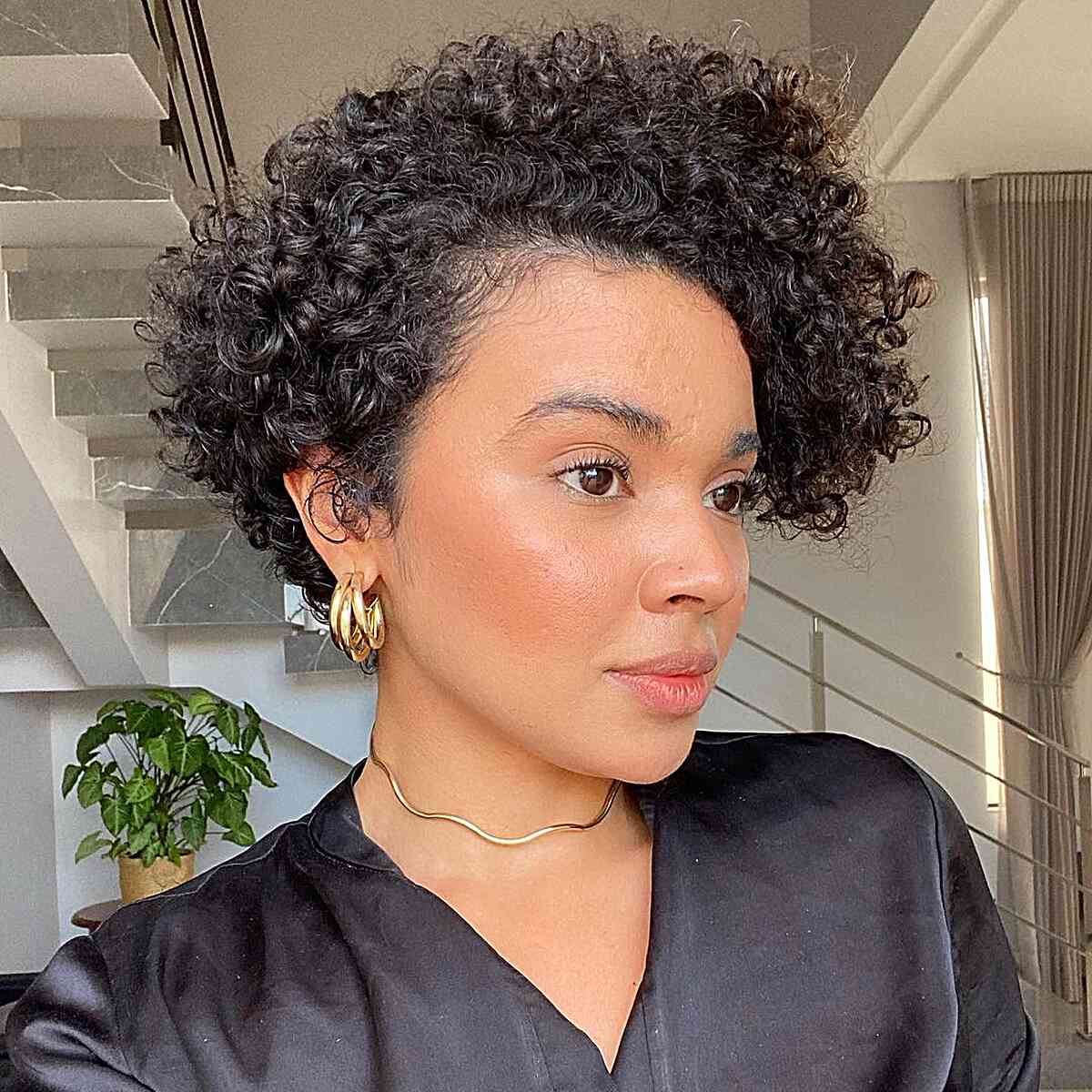 A woman with a square-shaped face wearing a dark curly pixie haircut, with rounded top curls that soften her jawline