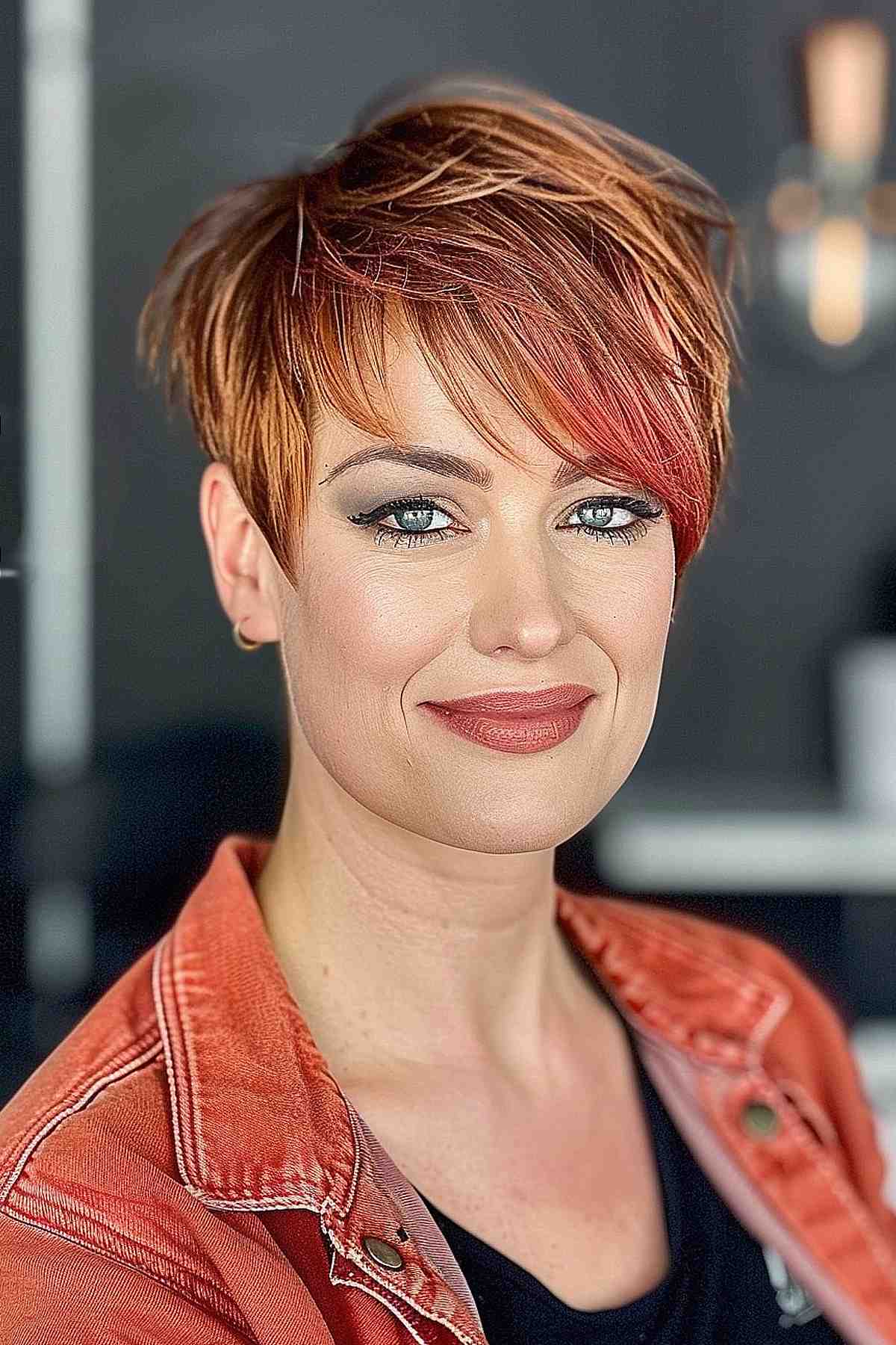 Woman with a playful pixie cut and asymmetric wispy bangs in a warm auburn color, looking stylish and approachable.